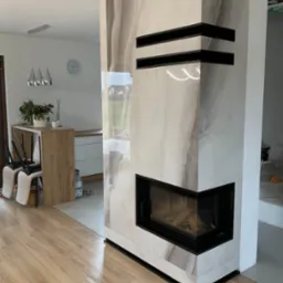 Nowoczesna obudowa paleniska z płyt imitujących marmur, czarne akcenty, widoczne drewno wewnątrz, fragment wnętrza z drewnianą podłogą i elementami wyposażenia.