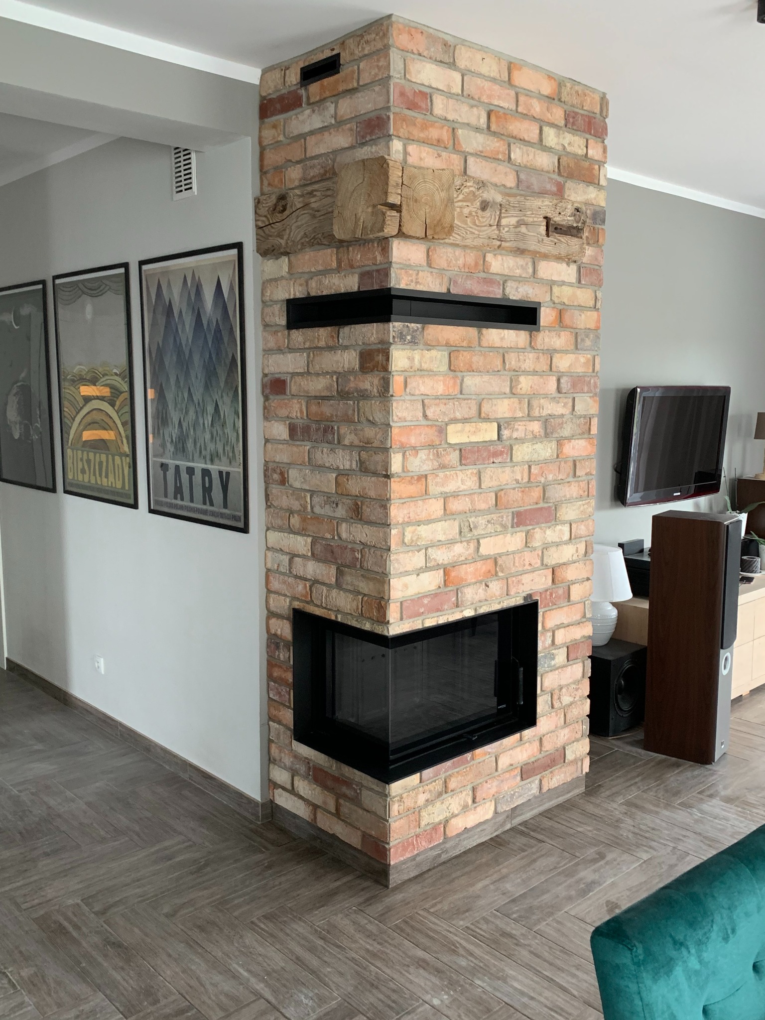Murowana konstrukcja z cegły z czarnym wkładem grzewczym, drewnianymi belkami i otworami wentylacyjnymi, w nowoczesnym salonie z szarymi ścianami i plakatami Bieszczady i Tatry.