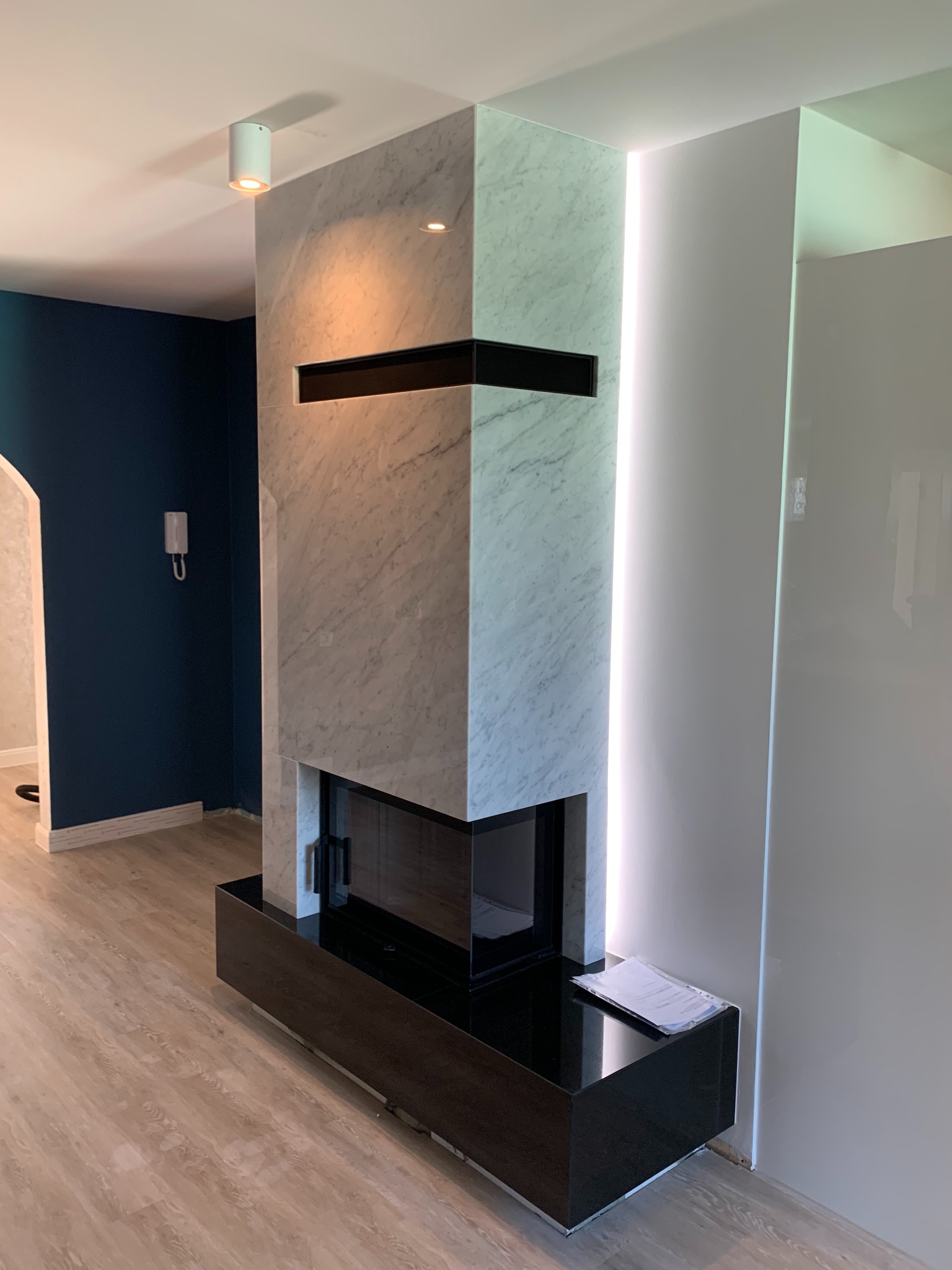 Nowoczesny kominek z marmurową obudową, czarnym wkładem i drewnopodobną podstawą, oświetlony punktowo i listwą LED, z dokumentami leżącymi obok, na tle granatowej ściany.