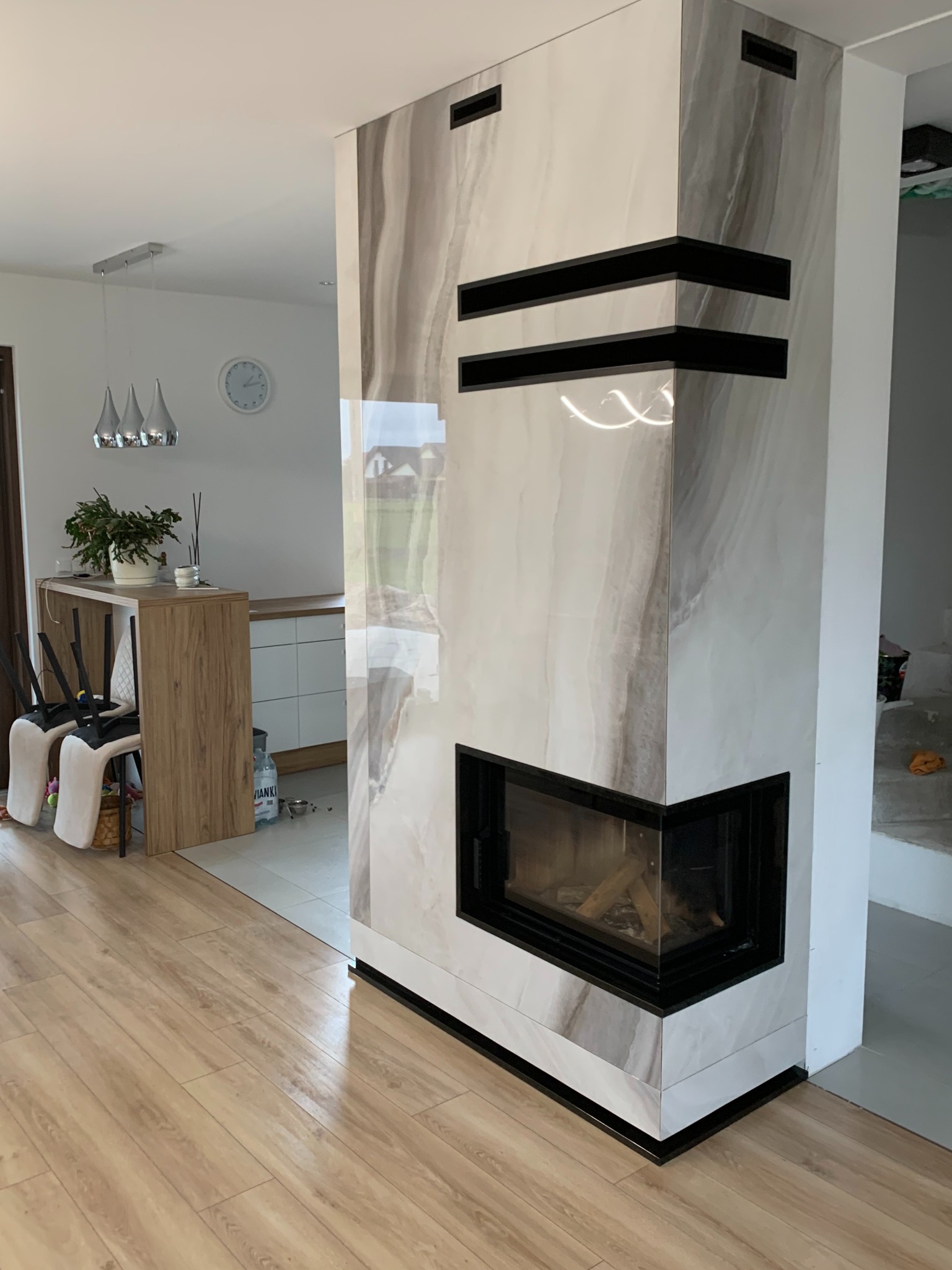 Nowoczesna obudowa paleniska z płyt imitujących marmur, czarne akcenty, widoczne drewno wewnątrz, fragment wnętrza z drewnianą podłogą i elementami wyposażenia.