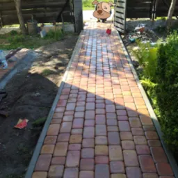 Ułożona kostka brukowa tworząca ścieżkę w ogrodzie, widoczne narzędzia i materiały budowlane w tle.