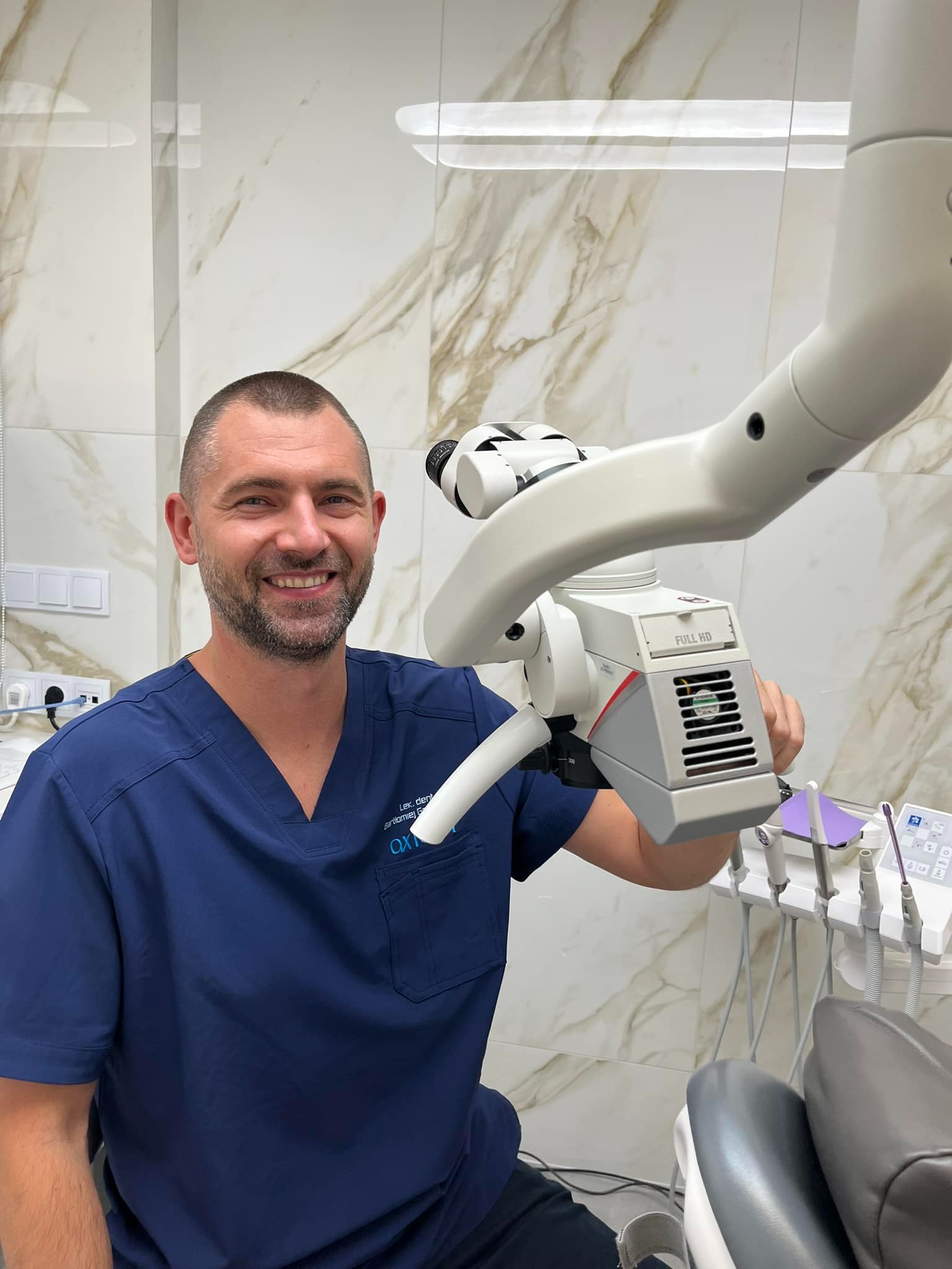 Uśmiechnięty stomatolog w granatowym uniformie, prezentujący mikroskop stomatologiczny Full HD w nowoczesnym gabinecie z marmurowymi płytkami ściennymi.