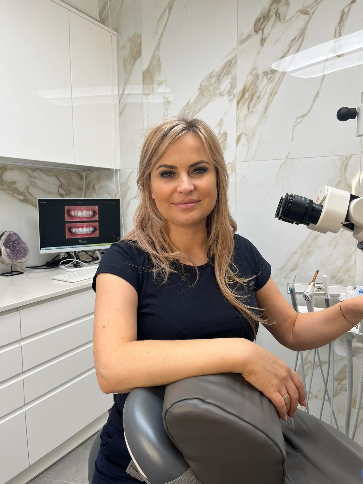 Uśmiechnięta kobieta w gabinecie, na monitorze widoczne zdjęcia zębów przed i po zabiegu, obok fotel dentystyczny i mikroskop.