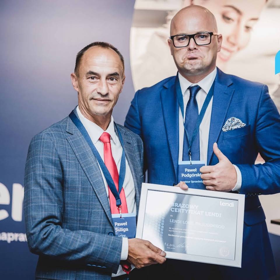 Dwóch mężczyzn w garniturach prezentuje certyfikat Lendi, jeden z nich z uniesionym kciukiem, obaj z identyfikatorami na szyi.