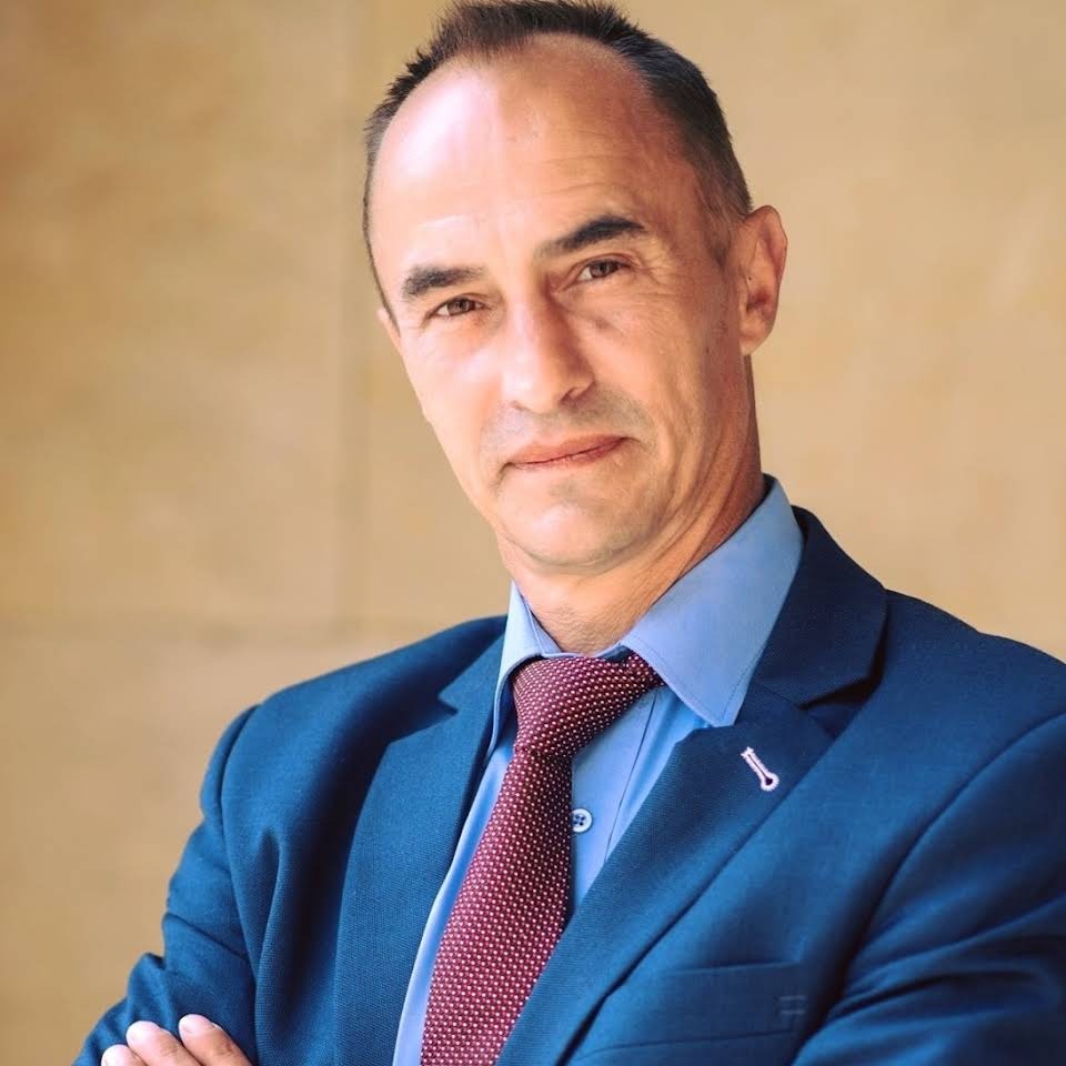 Portret mężczyzny w niebieskim garniturze i bordowym krawacie w kropki, z rękami skrzyżowanymi na piersiach i przypinką w klapie.