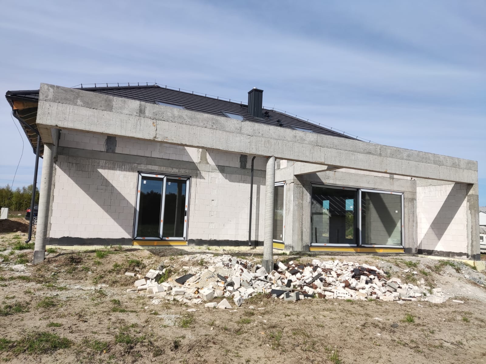 Surowy stan domu z betonu komórkowego z widocznymi belkami i słupami konstrukcyjnymi, czarną dachówką i gruzem na ziemi przed budynkiem.