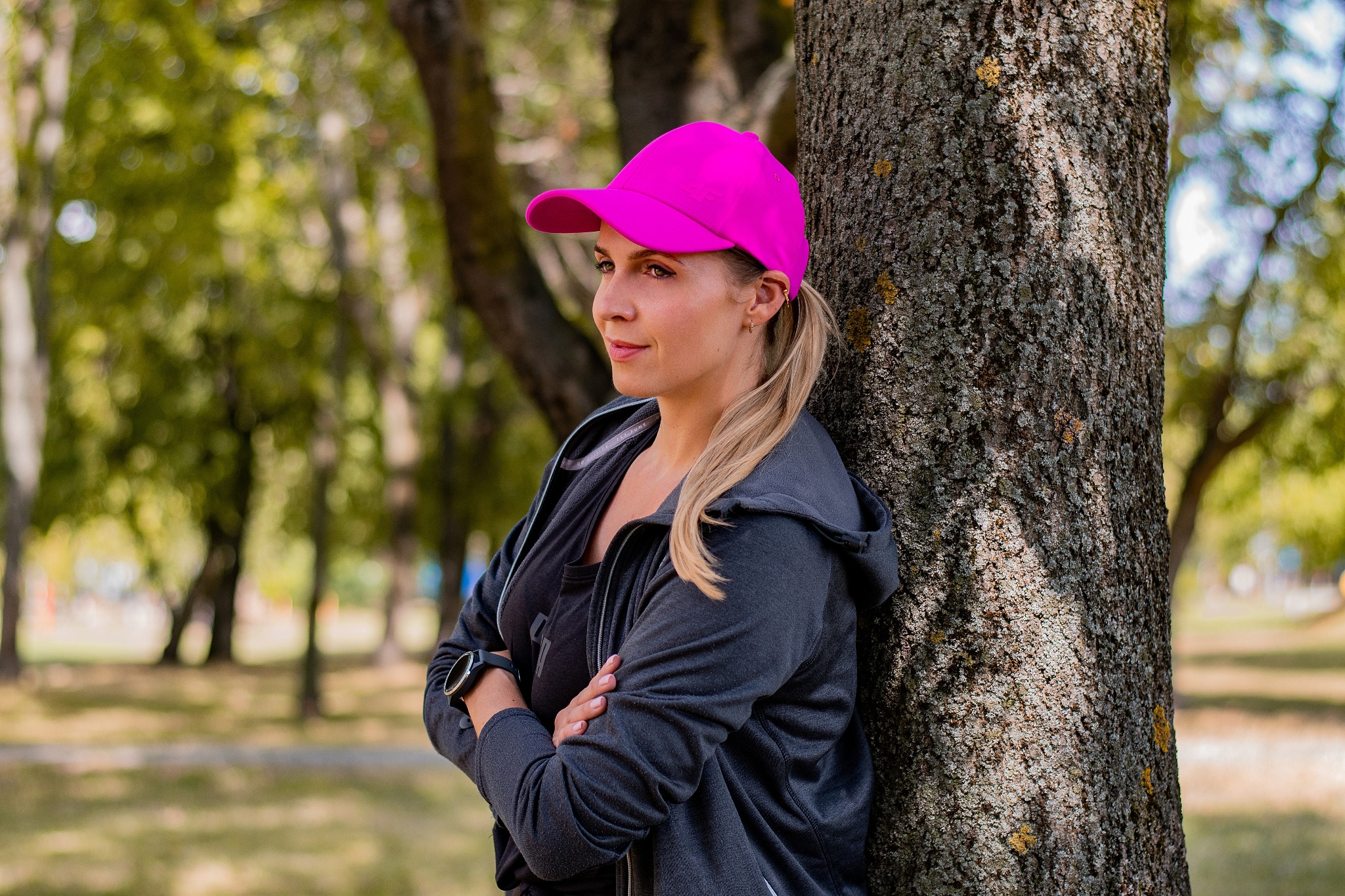 Kobieta w sportowym stroju i różowej czapce oparta o drzewo, z założonymi rękami i zegarkiem na nadgarstku, spogląda w dal.