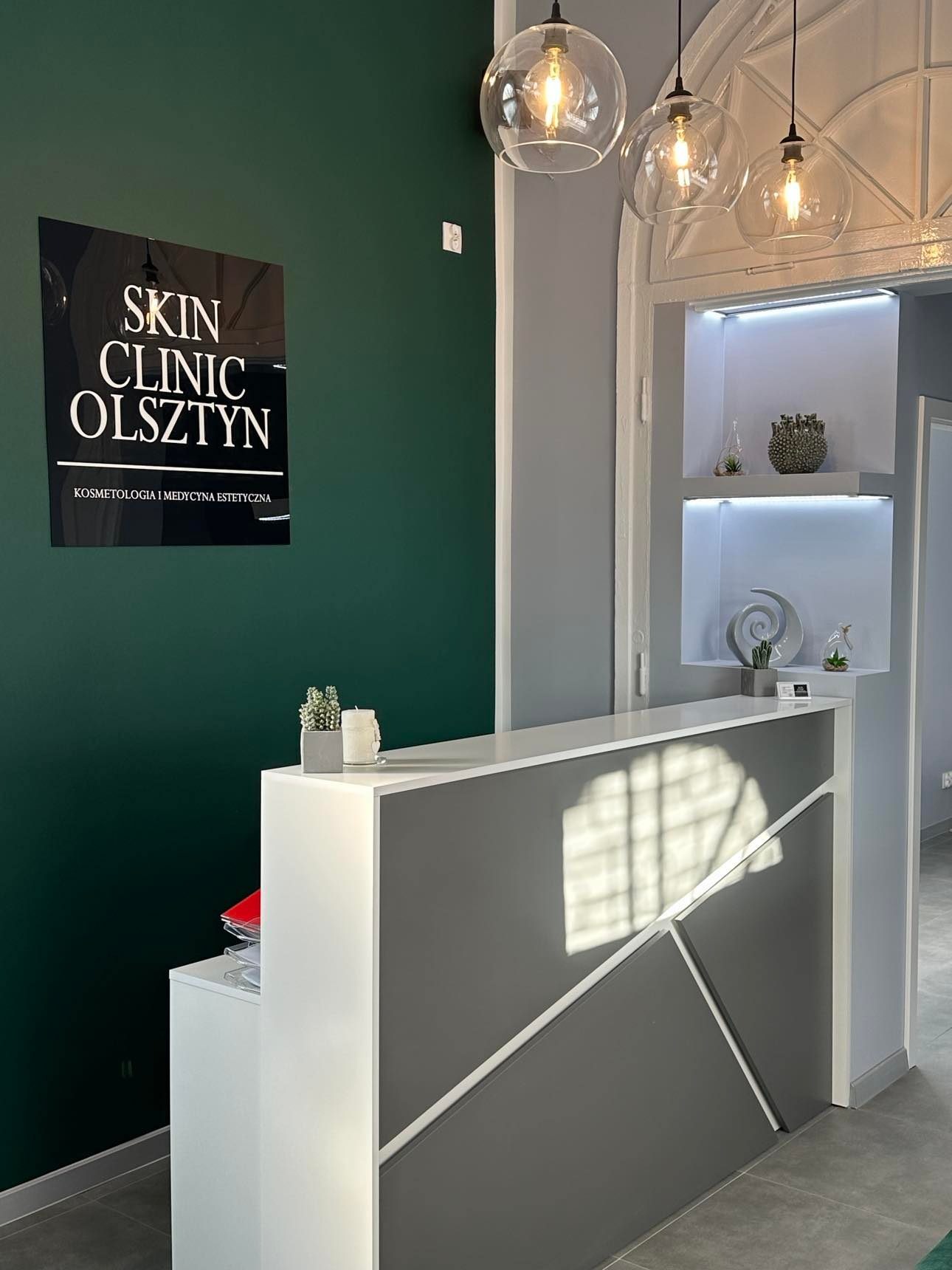 Recepcja Skin Clinic Olsztyn z ciemnozieloną ścianą, logo na czarnej tablicy, szaro-białym biurkiem recepcyjnym z geometrycznym wzorem, trzema szklanymi lampami wiszącymi i dekoracyjnymi półkami...