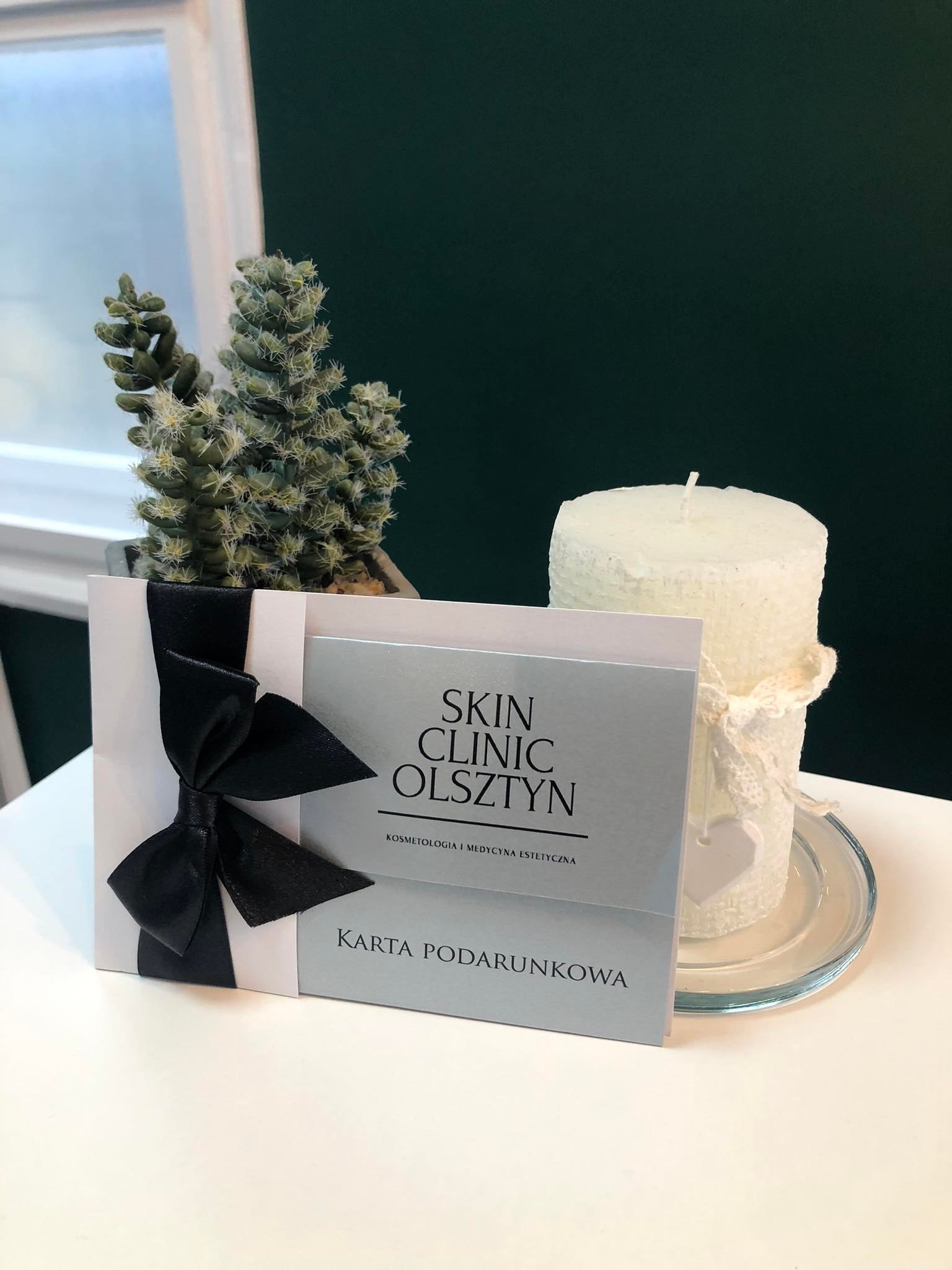 Karta podarunkowa Skin Clinic Olsztyn z czarną wstążką, kaktus w doniczce i biała świeca na szklanym talerzyku na białym stole, z zielonym tłem.