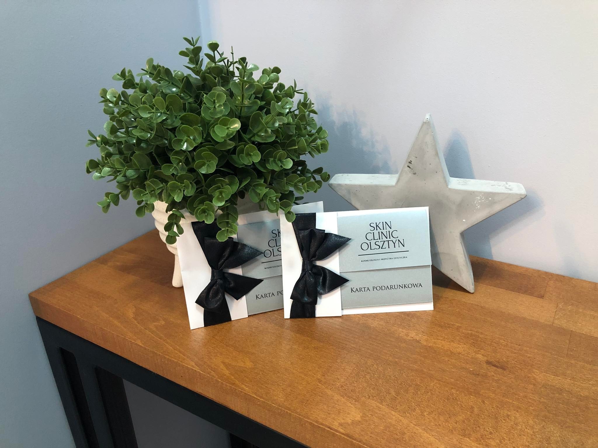 Dwie karty podarunkowe Skin Clinic Olsztyn z czarnymi kokardami na drewnianym stoliku obok rośliny w doniczce i betonowej gwiazdy.