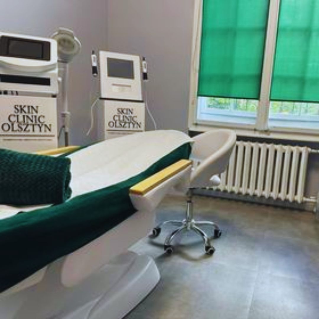 Klimatyczne wnętrze gabinetu z białym fotelem zabiegowym z zielonymi akcentami, obok sprzęt kosmetyczny z logo Skin Clinic Olsztyn, białe krzesło na kółkach i grzejnik pod oknem z zielonymi roletami.