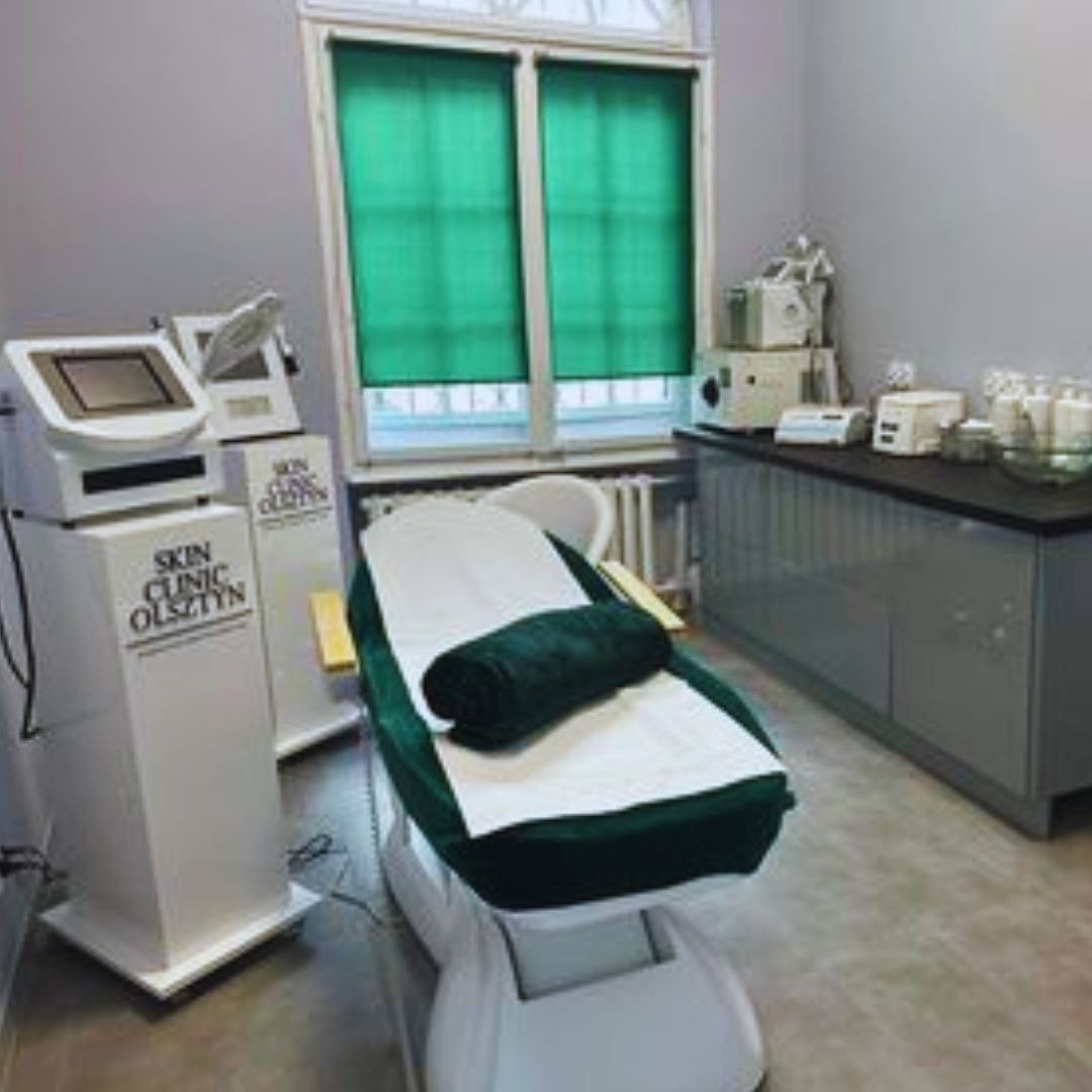Wnętrze gabinetu z fotelem kosmetycznym z zielonym obiciem i ręcznikiem, obok białe urządzenia z logo 'Skin Clinic Olsztyn', w tle okno z zielonymi roletami.