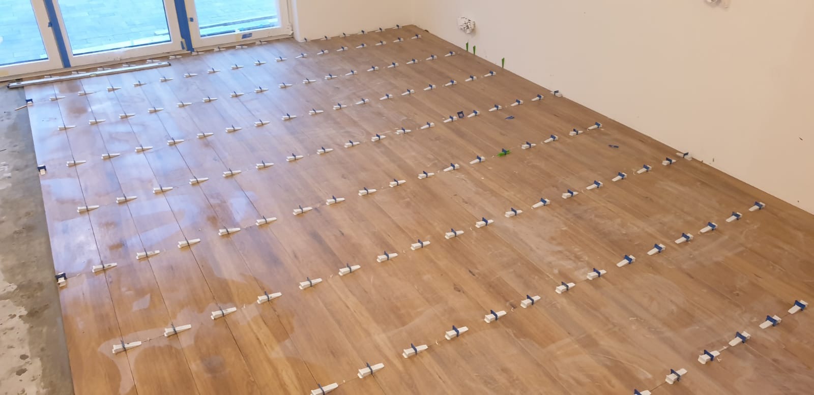 Układanie paneli podłogowych w jasnym pomieszczeniu z widocznymi białymi klipsami poziomującymi i niebieskimi klinami, duże okno w tle.