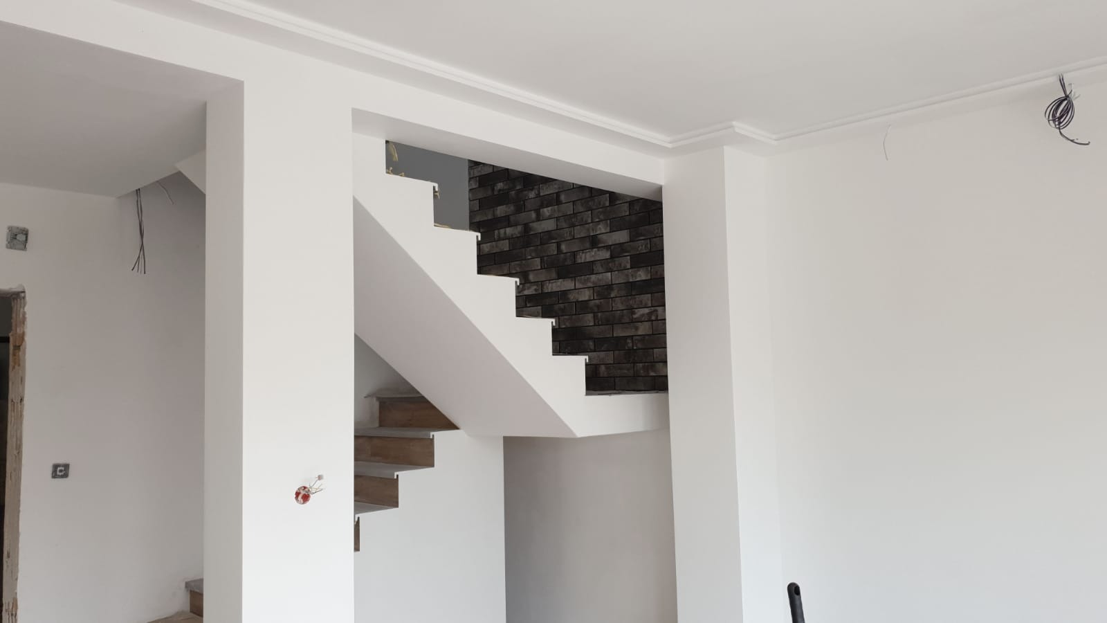 Wnętrze w trakcie prac wykończeniowych: białe ściany z listwami przysufitowymi, schody z drewnianymi stopniami i fragment ściany wyłożonej ciemną cegłą, widoczne przewody elektryczne.
