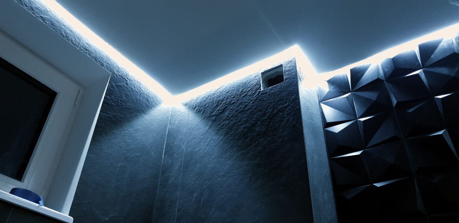 Nowoczesna łazienka z oświetleniem LED podsufitowym, ciemnymi płytkami imitującymi kamień i trójwymiarową ścianą dekoracyjną.