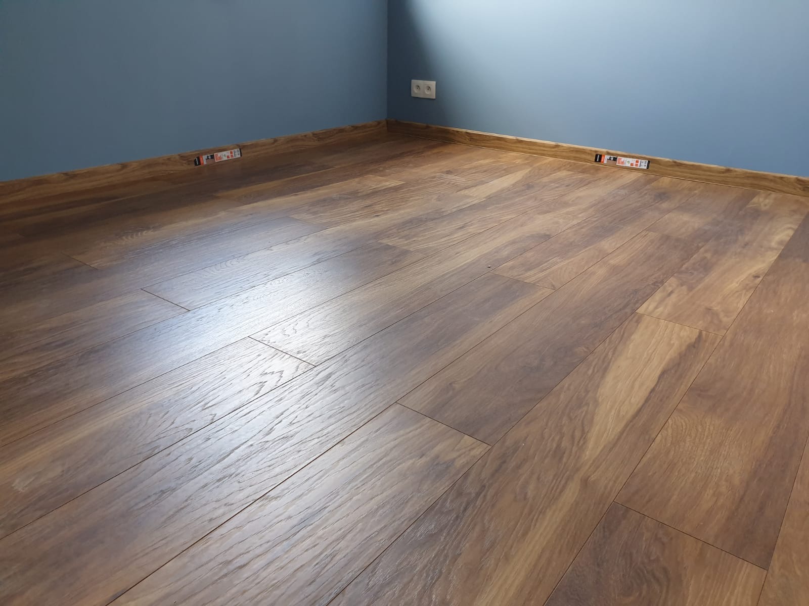 Ukończone układanie paneli podłogowych w rogu pokoju z niebieskimi ścianami, widoczne listwy przypodłogowe i gniazdko elektryczne.