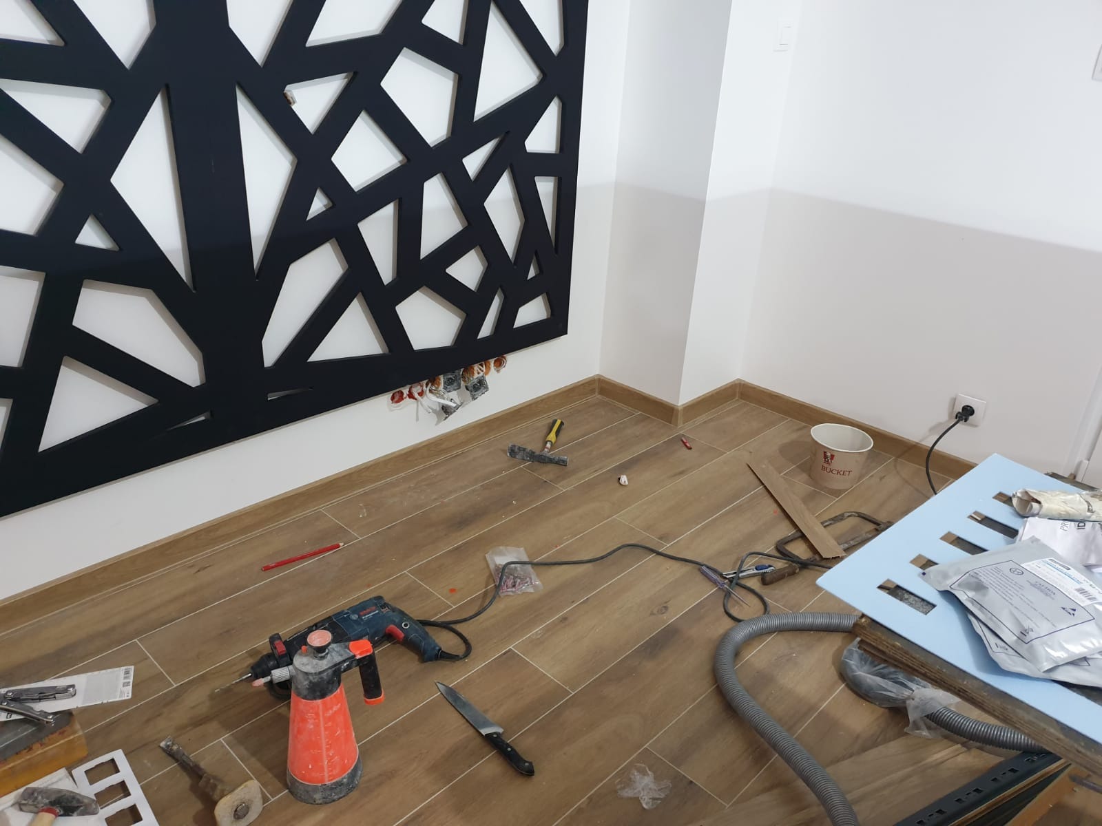 Montaż dekoracyjnej, czarnej, ażurowej płyty ściennej w trakcie prac wykończeniowych. Widoczne narzędzia, przewody elektryczne i fragmenty płytek podłogowych imitujących drewno.