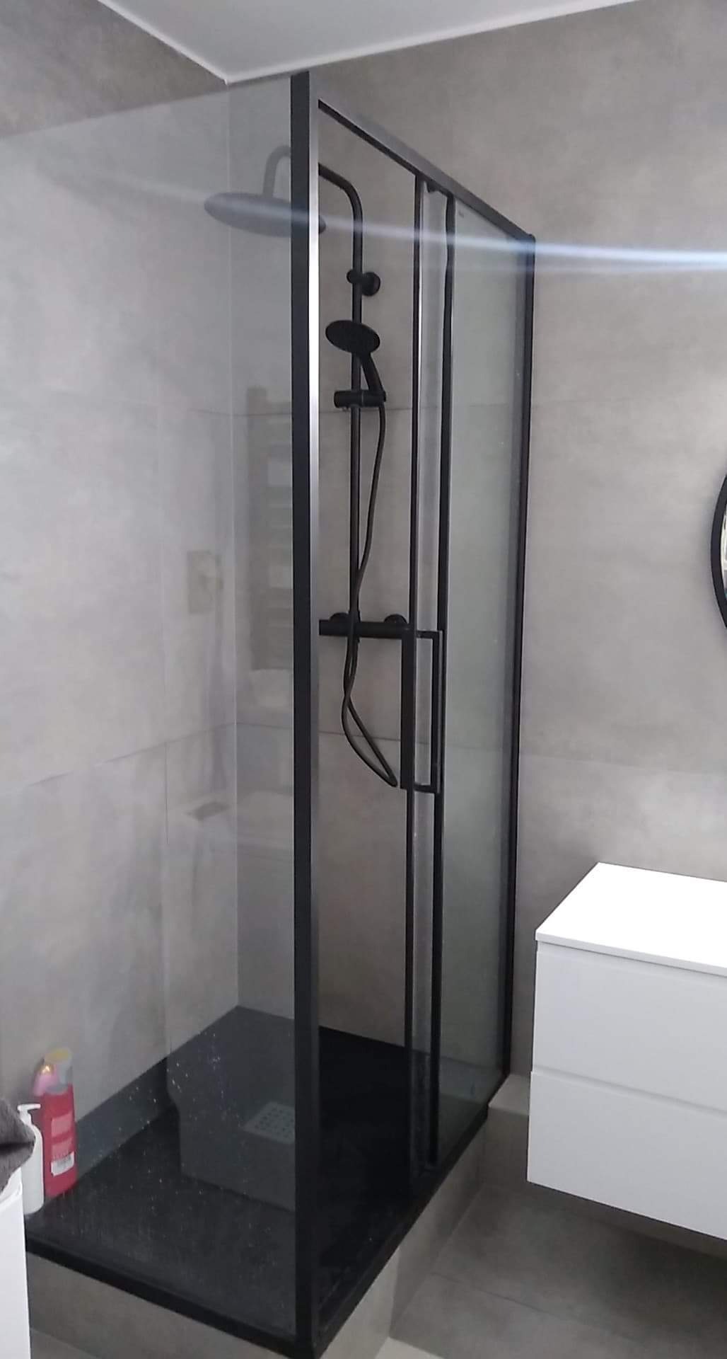 Nowoczesna kabina prysznicowa z czarnymi profilami i deszczownicą, czarny brodzik z delikatnym połyskiem, szare płytki na ścianach, minimalistyczna biała szafka umywalkowa.