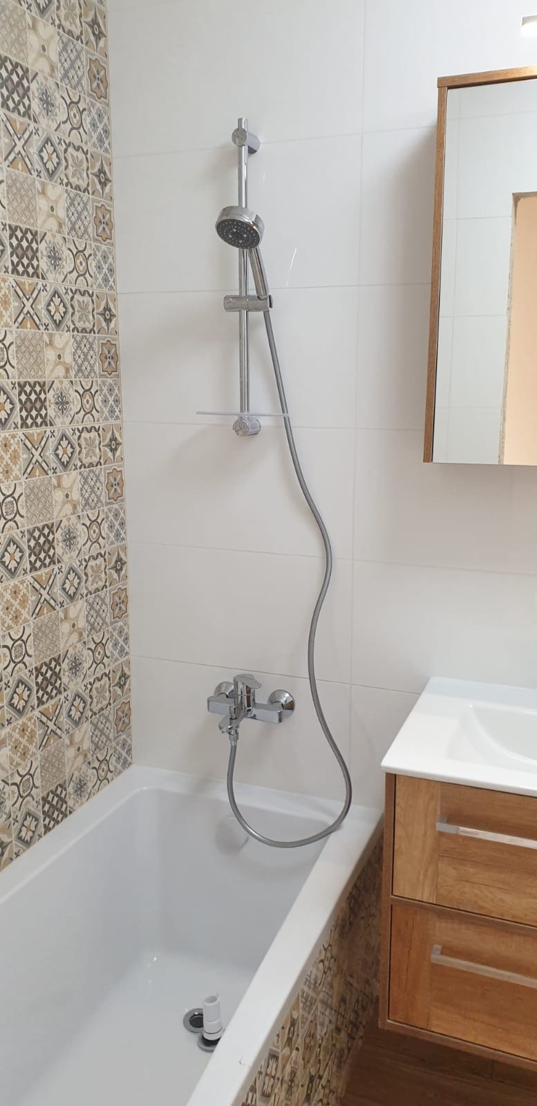 Wykończona łazienka z wanną, chromowaną armaturą natryskową i umywalką w szafce z drewnopodobnym fornirem, dekoracyjne płytki patchwork na ścianie.