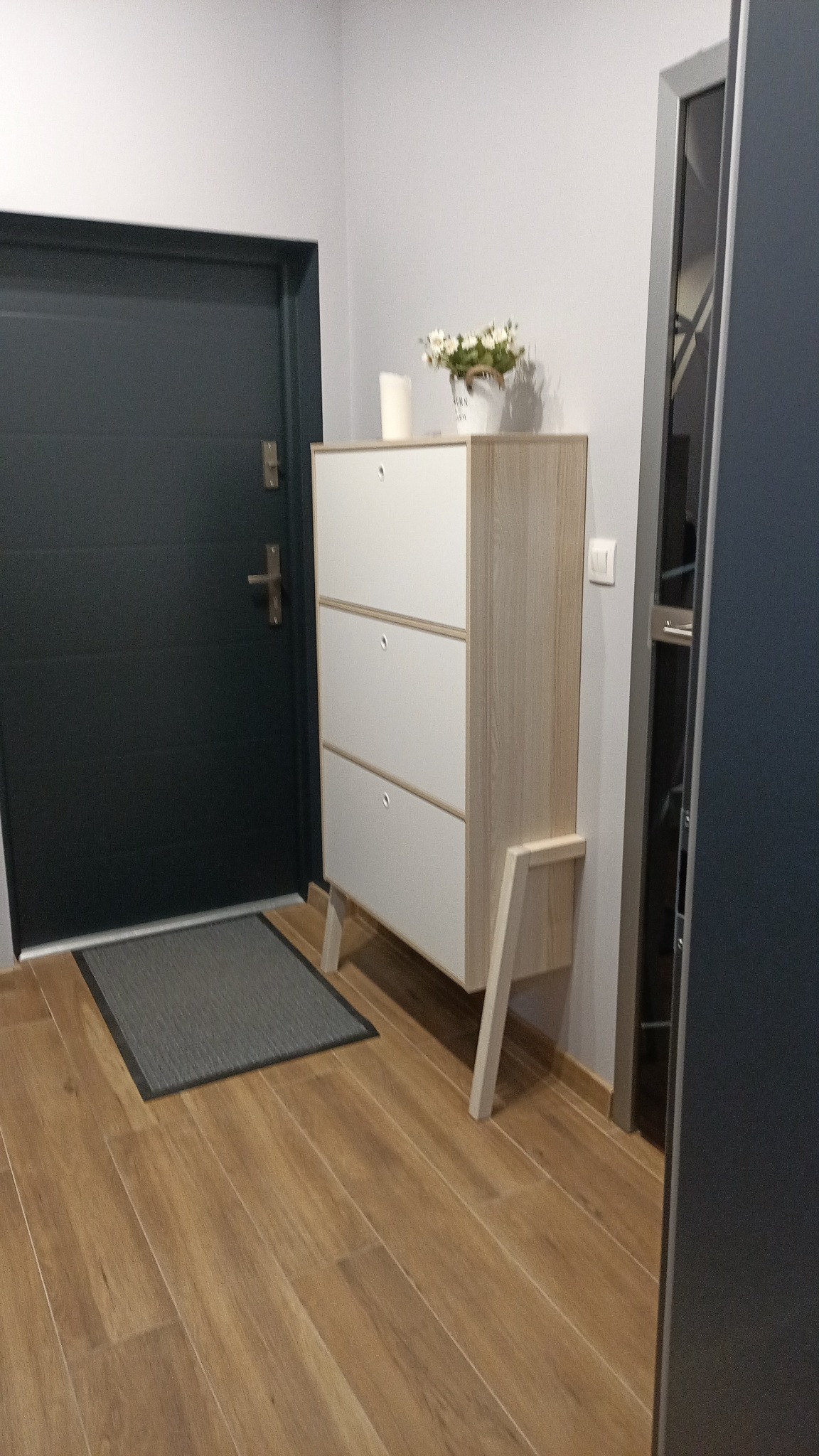 Nowoczesny przedpokój z drewnianą podłogą, szarą wycieraczką, szafką na buty z trzema frontami i dekoracją kwiatową, ciemne drzwi wejściowe.