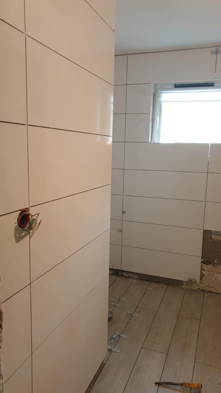 Białe, poziome płytki ścienne z widoczną puszką elektryczną i wystającymi przewodami, w tle okno i częściowo ułożona podłoga z płytek, widoczne krzyżyki dystansowe.