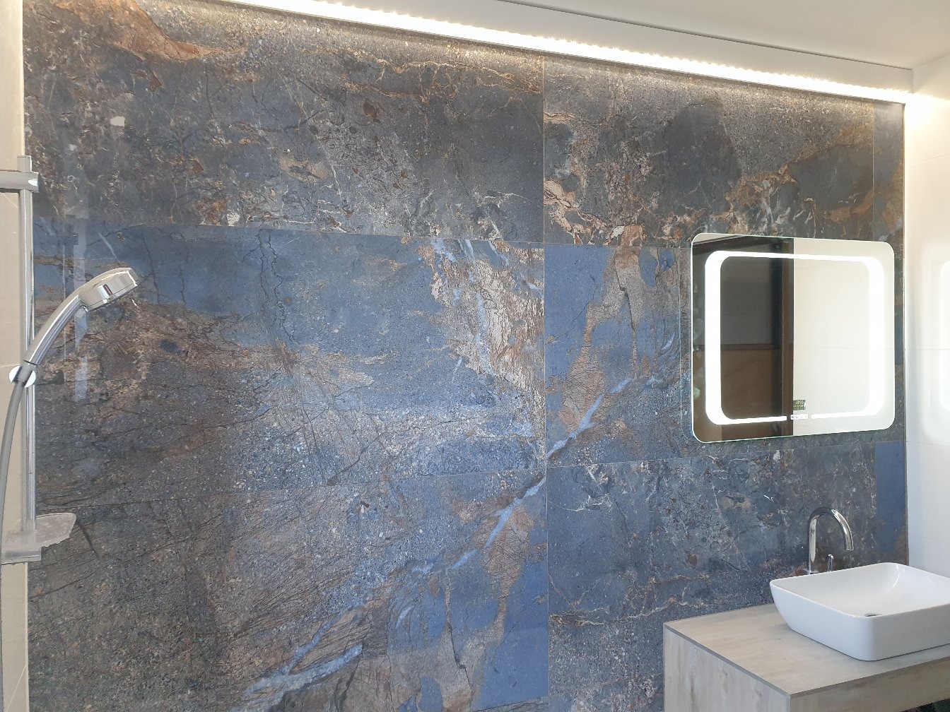 Nowoczesna łazienka z płytkami imitującymi marmur w odcieniach szarości i niebieskiego, podświetlane lustro LED w kształcie kwadratu z zaokrąglonymi rogami oraz biała, prostokątna umywalka nablatowa.