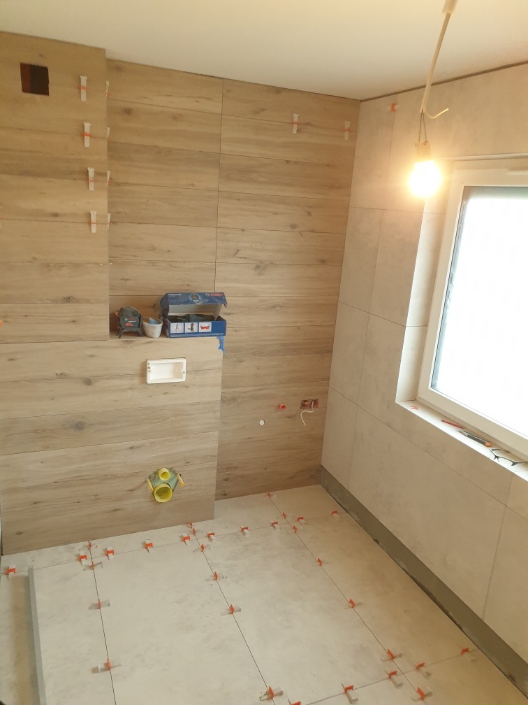 Montaż płytek imitujących drewno na ścianach i podłodze w łazience, widoczne krzyżyki dystansowe, pudełko po laserze krzyżowym Bosch, żółta rura kanalizacyjna w ścianie.