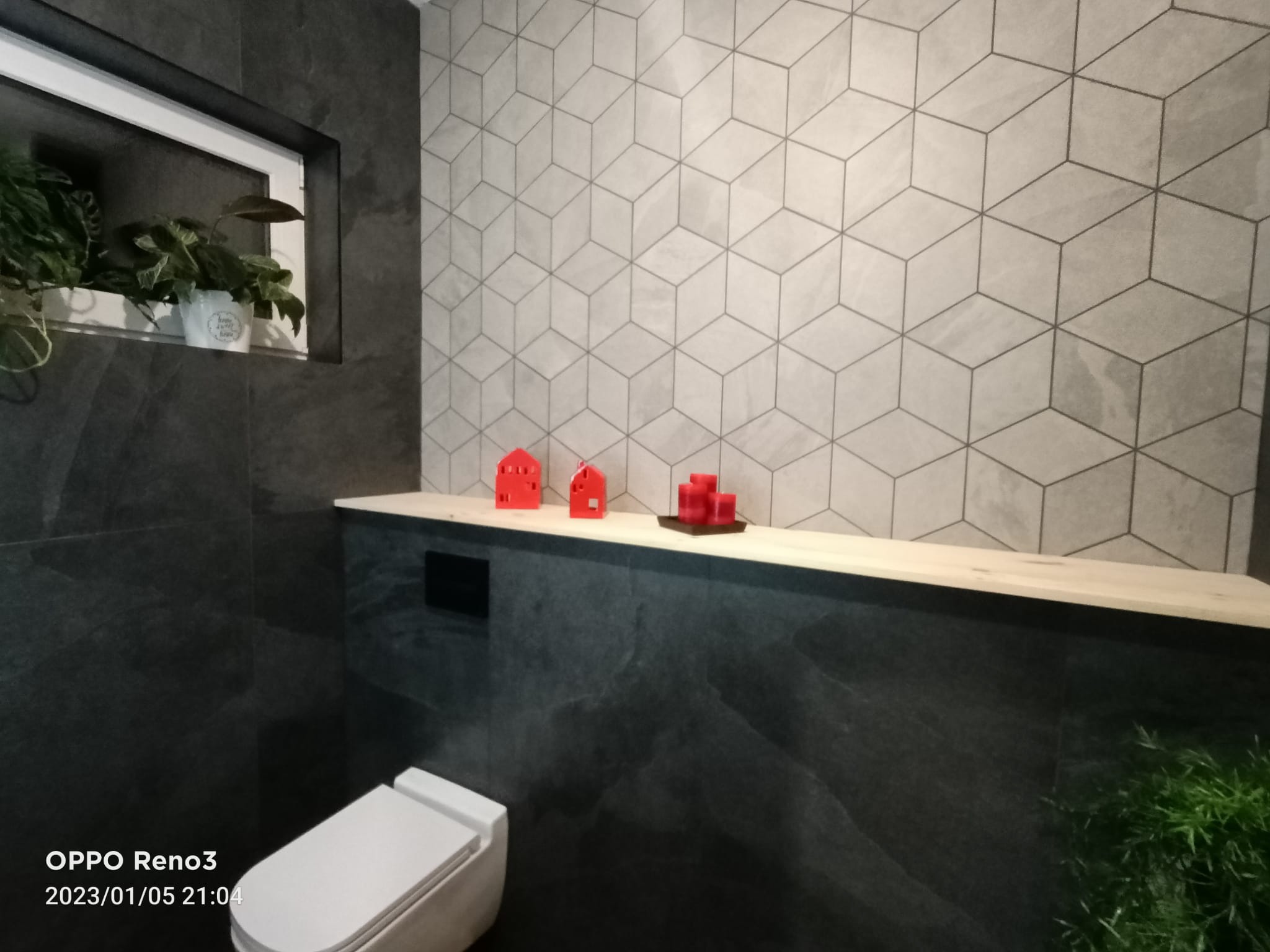 Nowoczesna łazienka z ciemnymi, marmurkowymi płytkami, geometryczną tapetą w odcieniach szarości, drewnianą półką z dekoracjami: czerwone domki i świeczki, oraz roślinami w doniczkach w niszy i...