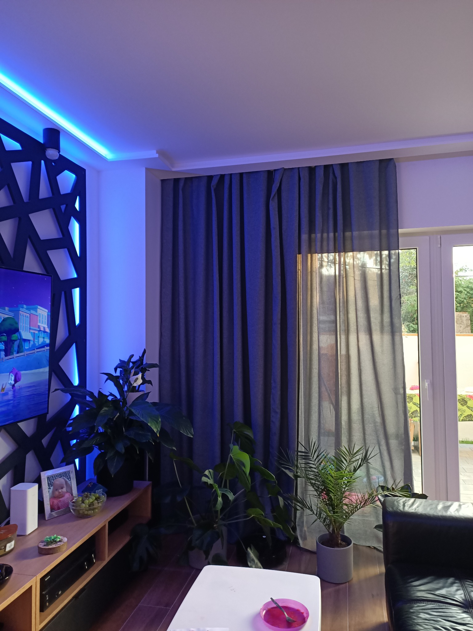 Nowoczesny salon z dekoracyjną ścianą podświetloną niebieskim LED, szare zasłony przy oknie balkonowym i rośliny doniczkowe na drewnianej komodzie.