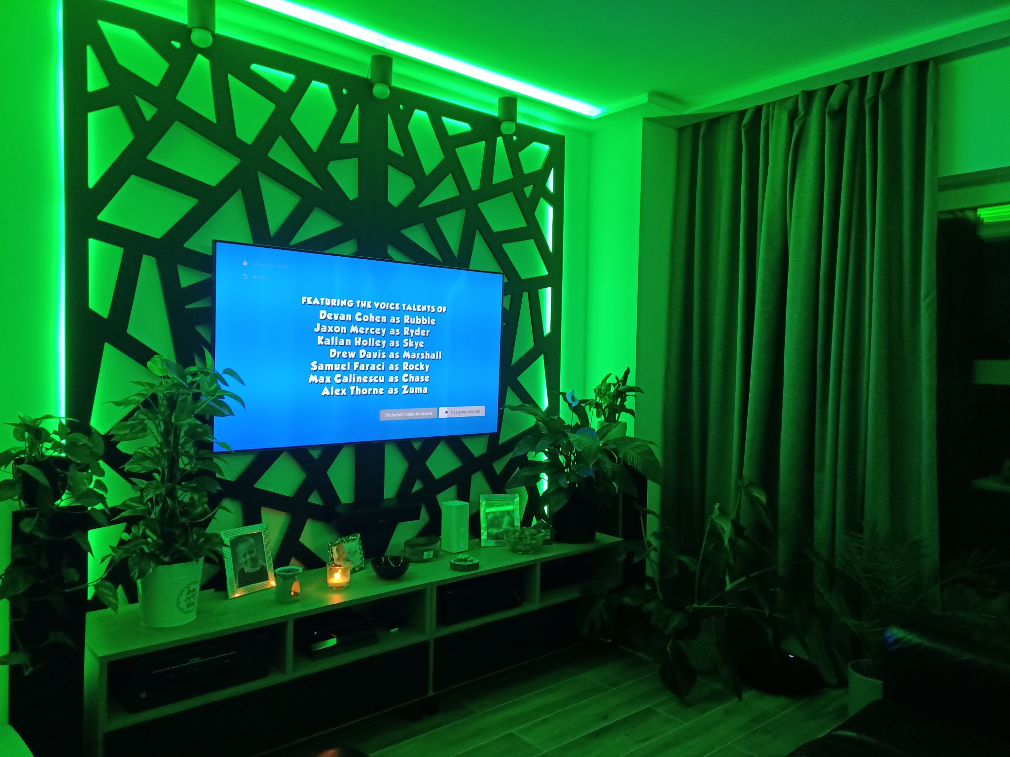 Salon z podświetloną na zielono dekoracyjną ścianą z geometrycznym wzorem, telewizorem wyświetlającym listę aktorów głosowych, roślinami doniczkowymi i drewnianą podłogą, wszystko utrzymane...