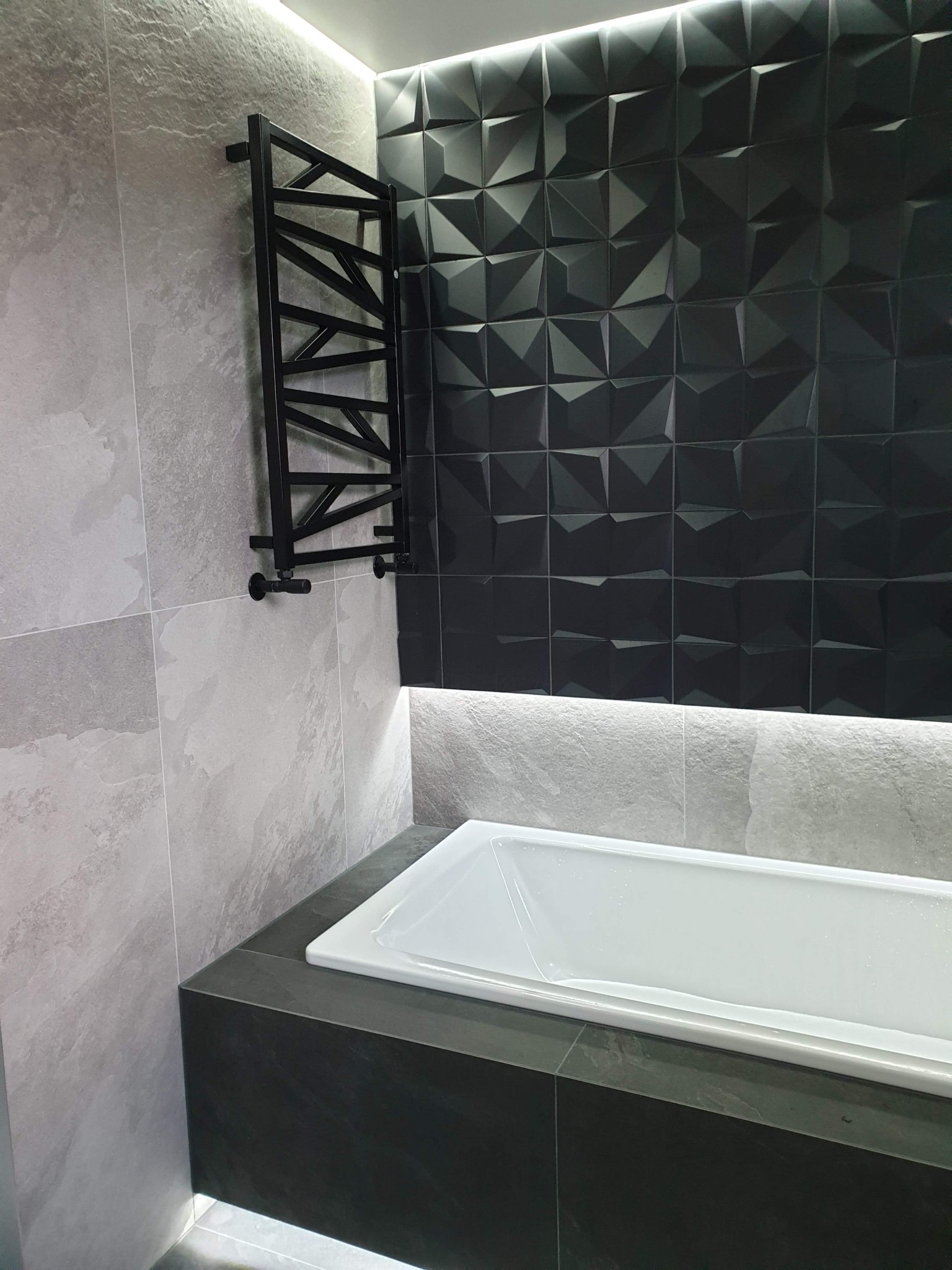 Nowoczesna łazienka z czarno-białą kolorystyką, ozdobną czarną ścianą 3D, grzejnikiem drabinkowym i podświetlaną wanną.