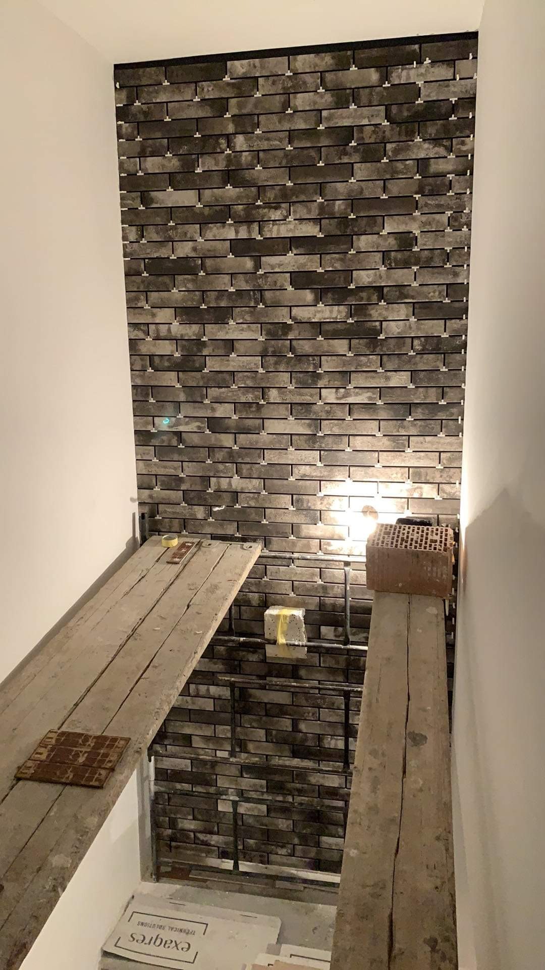 Montaż szarej, ceglanej okładziny ściennej z użyciem krzyżyków dystansowych, widoczne rusztowanie robocze i materiały budowlane.