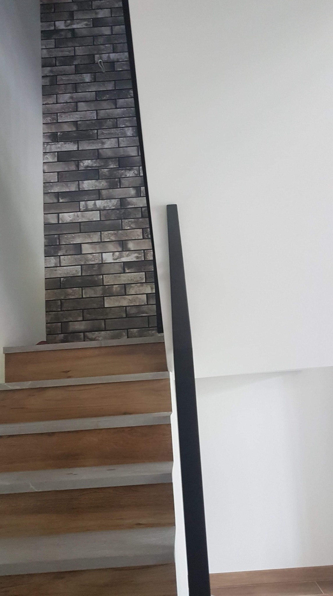 Schody wykończone panelami drewnopodobnymi, ściana z dekoracyjną okładziną ceglaną, biała ściana i czarna poręcz.