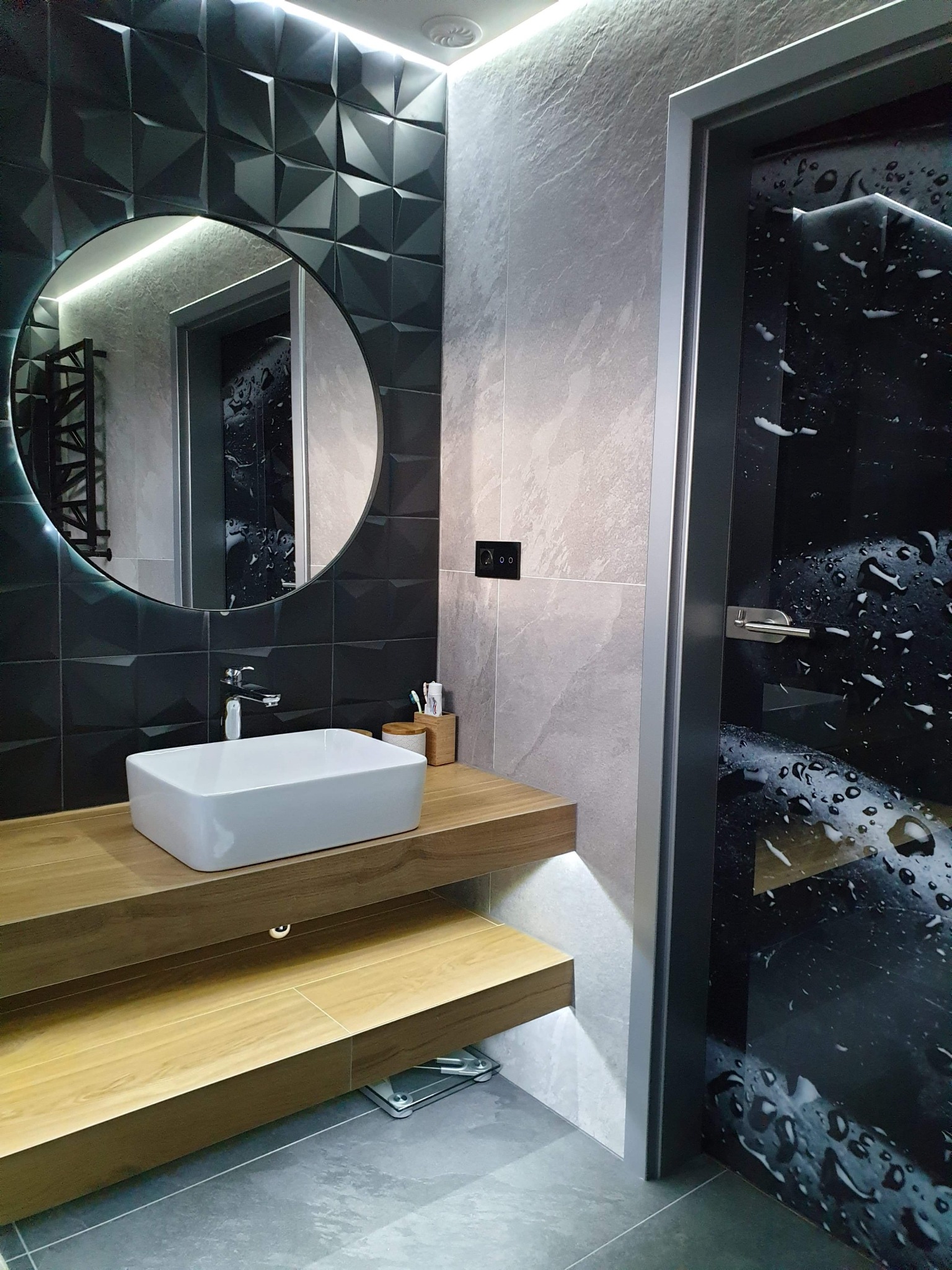 Nowoczesna łazienka z geometryczną czarną ścianą, okrągłym lustrem z podświetleniem LED, drewnianym blatem z białą umywalką i drzwiami z motywem kropli wody.