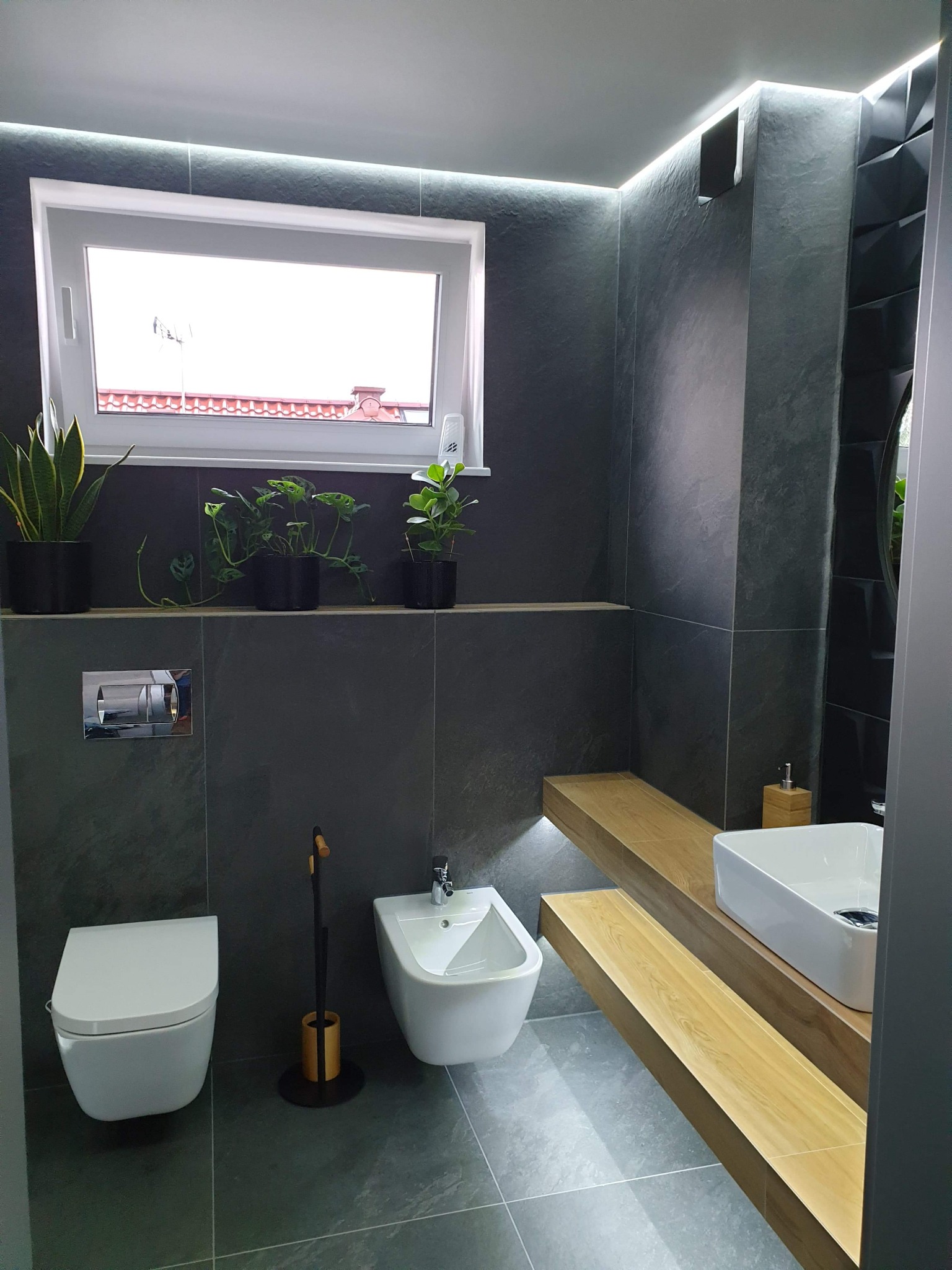 Nowoczesna łazienka z szarymi płytkami imitującymi kamień, podświetleniem LED, wiszącą toaletą i bidetem, drewnianymi półkami i roślinami w doniczkach.