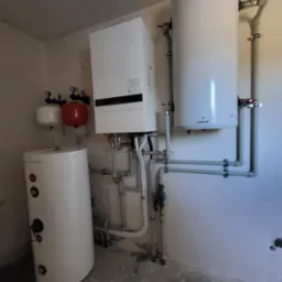 Zestaw urządzeń grzewczych w kotłowni: biały zbiornik Galmet, pionowy bojler, ekspansomat czerwony i biały, oraz rury z zaworami.