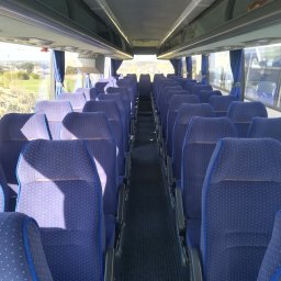 Auto Land - Wnętrze autobusu z rzędami niebieskich foteli z białymi kropkami, widok z przodu na puste przejście, zasłonięte okna po bokach i widok na krajobraz za oknem.