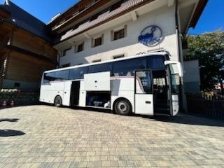 Biały autokar turystyczny z otwartymi lukami bagażowymi, zaparkowany przed budynkiem z logo firmy przewozowej, na tle brukowanej nawierzchni.