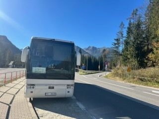 Biały autokar turystyczny zaparkowany na parkingu przy drodze z widokiem na góry w oddali, błękitne niebo.