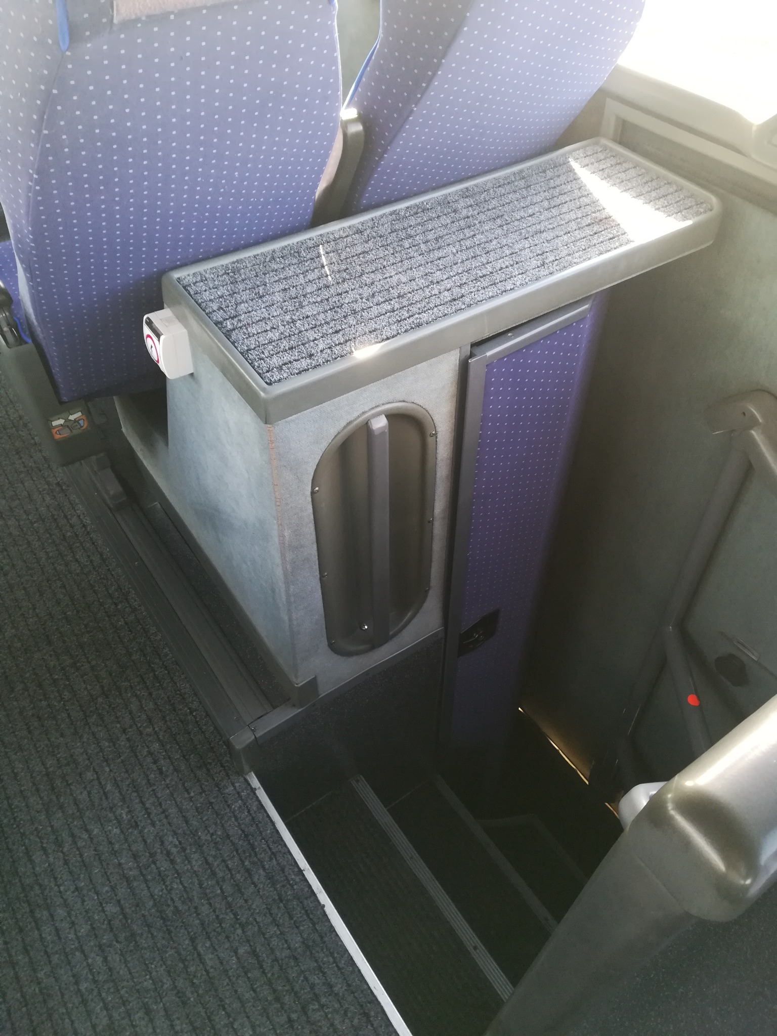 Wnętrze autobusu dalekobieżnego: schody wejściowe, poręcz, podłokietnik z szarą wykładziną, siedzenia z niebieską tapicerką w kropki.
