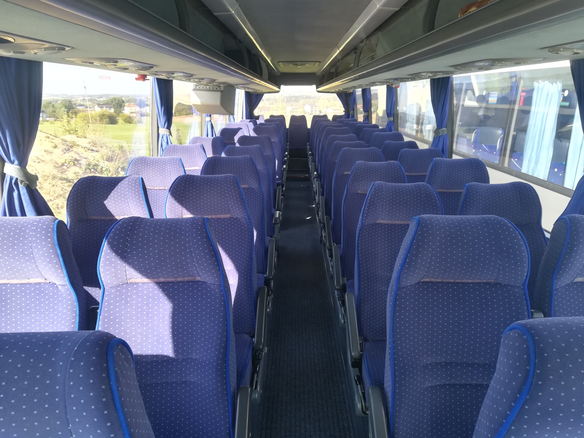 Wnętrze autobusu z rzędami niebieskich foteli z białymi kropkami, widok z przodu na puste przejście, zasłonięte okna po bokach i widok na krajobraz za oknem.
