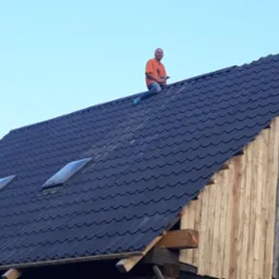Pracownik w pomarańczowej koszulce siedzi na nowym, ciemnym dachu z dwoma świetlikami, trzymając w ręku wkrętarkę; widoczne drewniane elementy konstrukcji budynku.