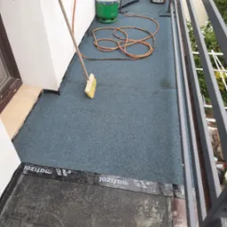 Balkon w trakcie uszczelniania: widoczna papa na podłożu, butla gazowa, wiadro z masą bitumiczną, wąż oraz szczotka do rozprowadzania preparatu.