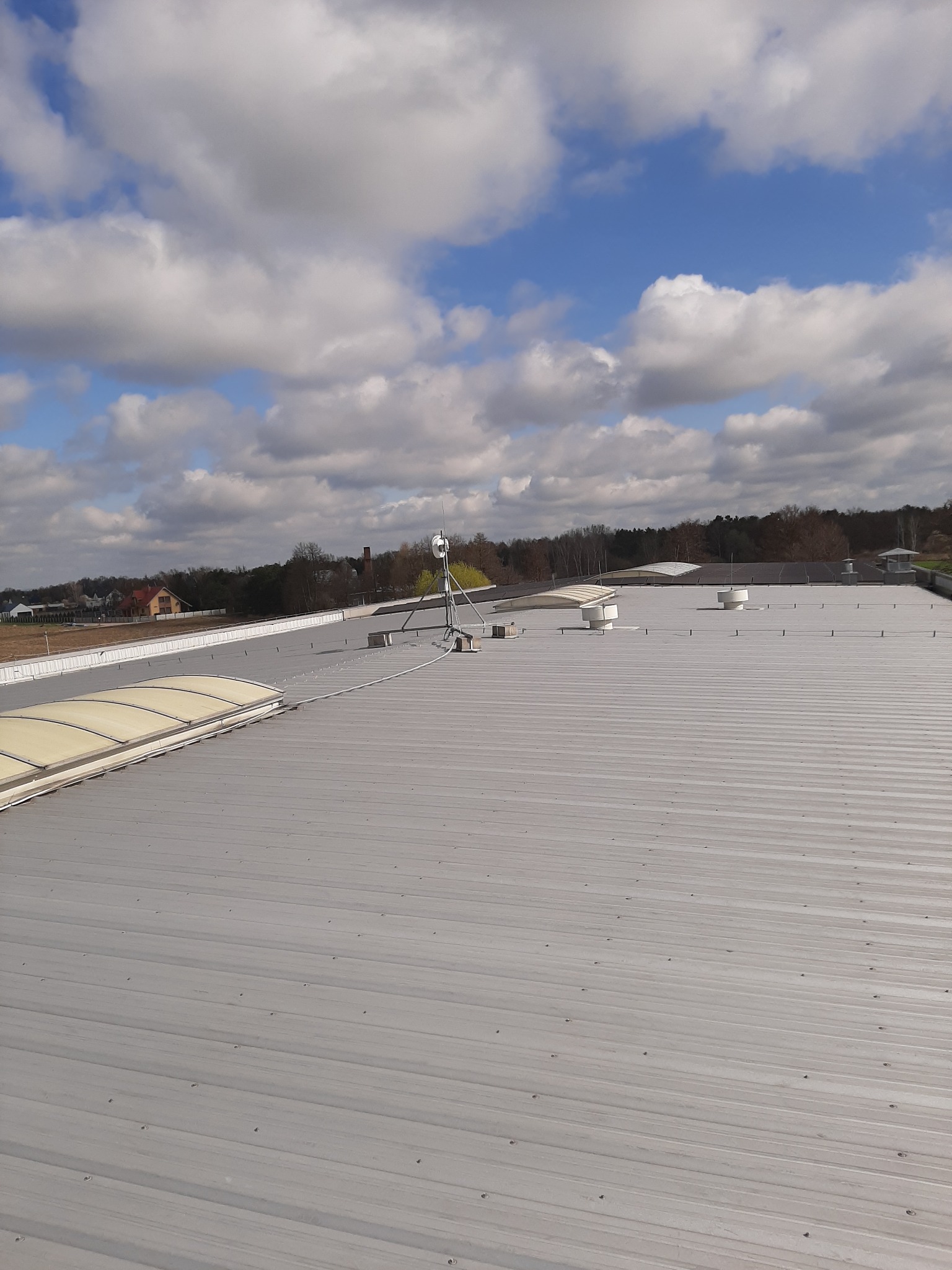 Szary dach z blachy trapezowej w Olszynie z widocznymi świetlikami dachowymi i anteną, na tle błękitnego nieba z chmurami.