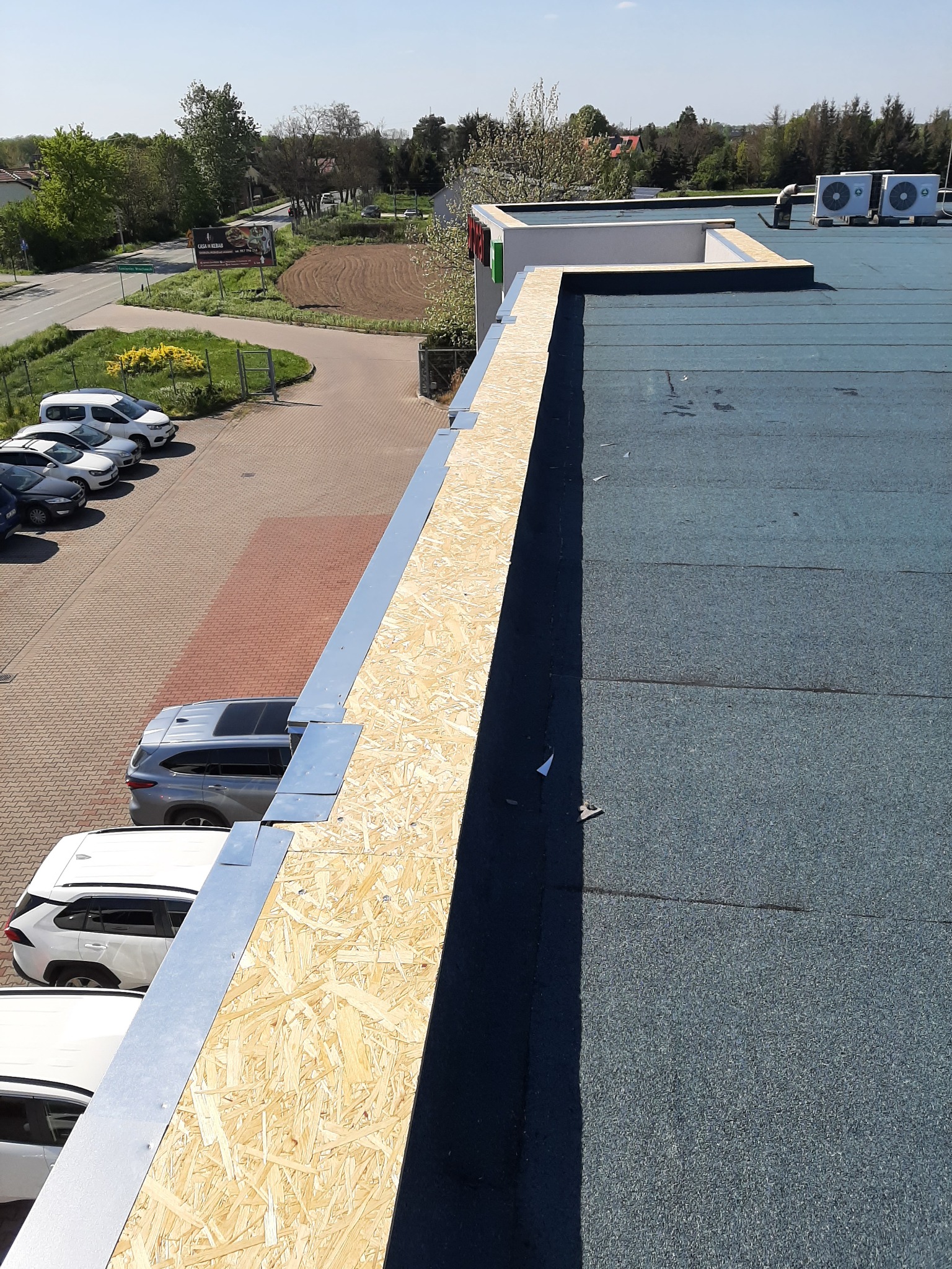 Płaski dach budynku komercyjnego pokryty papą, z zamontowaną obróbką blacharską i płytami OSB na krawędzi dachu, widok z góry na parking i okolicę.