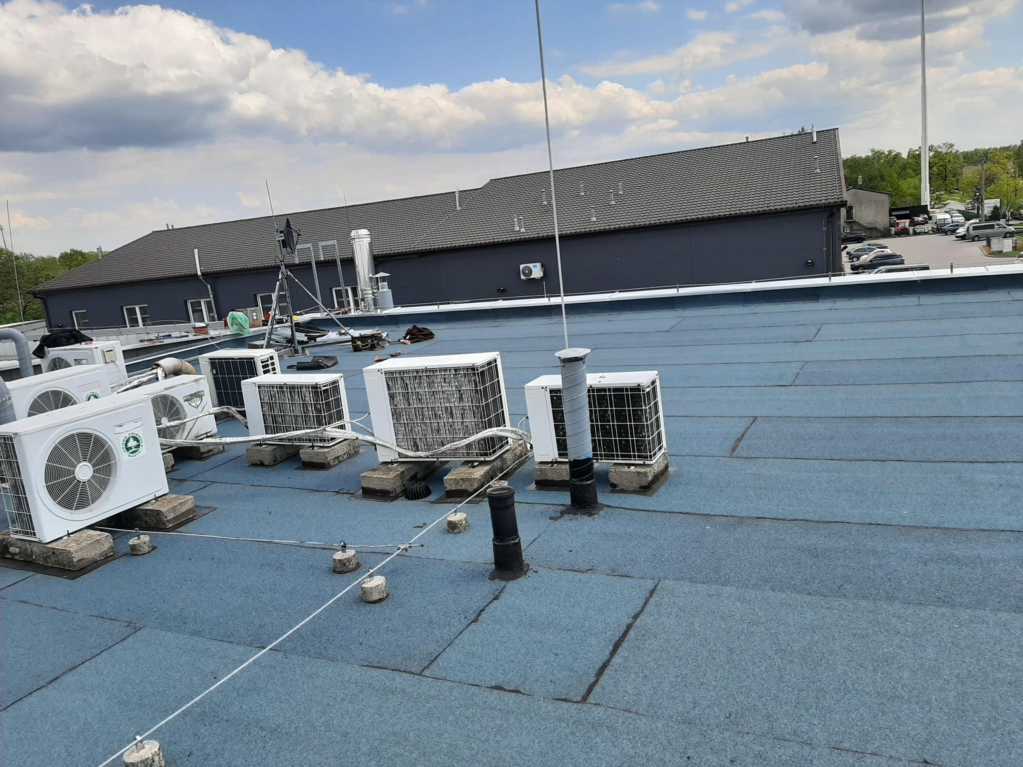 Płaski dach pokryty papą termozgrzewalną w kolorze niebieskim, na którym zamontowano liczne jednostki zewnętrzne klimatyzacji, zabezpieczone systemem odgromowym z przewodami i betonowymi...