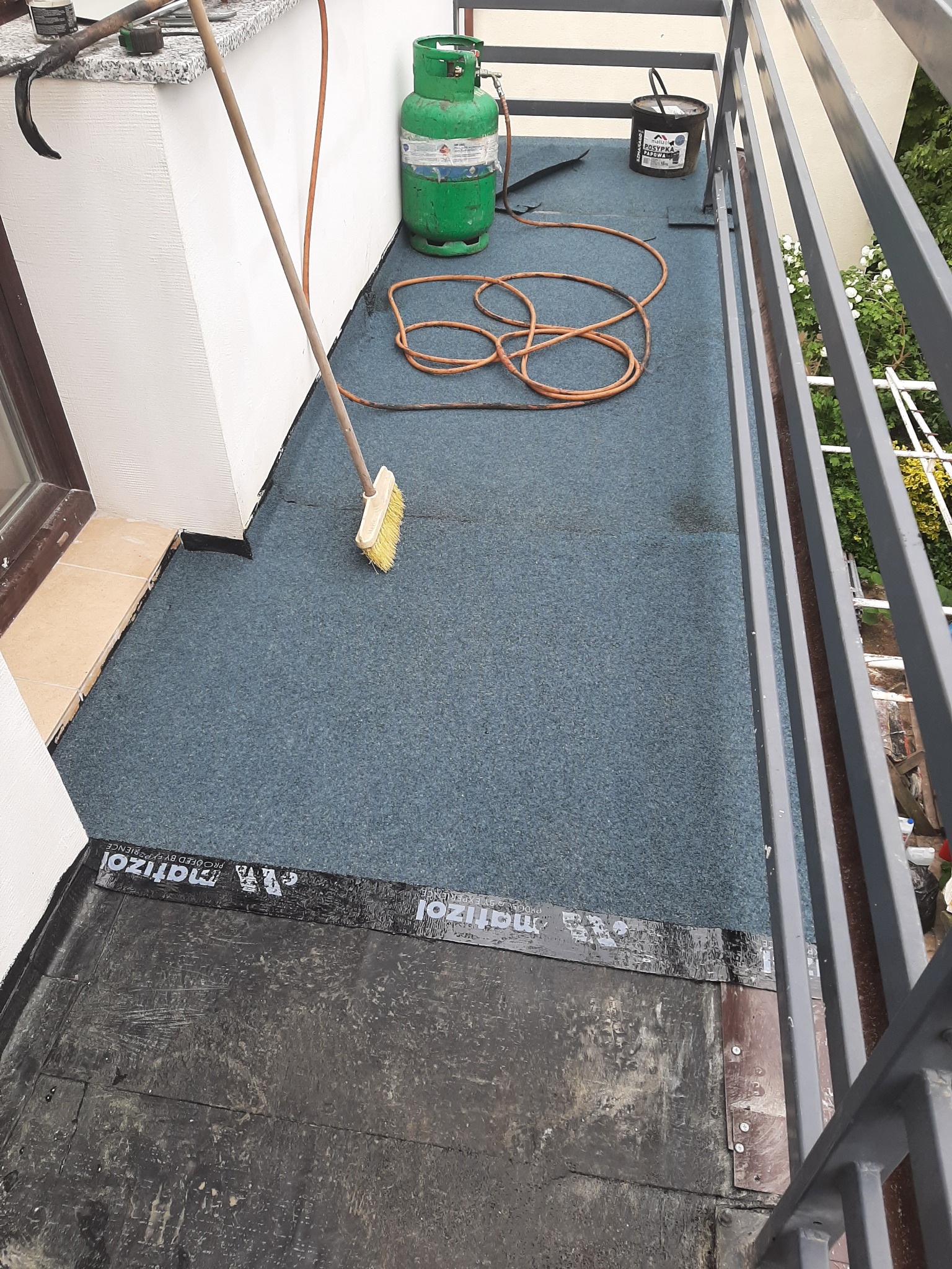 Balkon w trakcie uszczelniania: widoczna papa na podłożu, butla gazowa, wiadro z masą bitumiczną, wąż oraz szczotka do rozprowadzania preparatu.