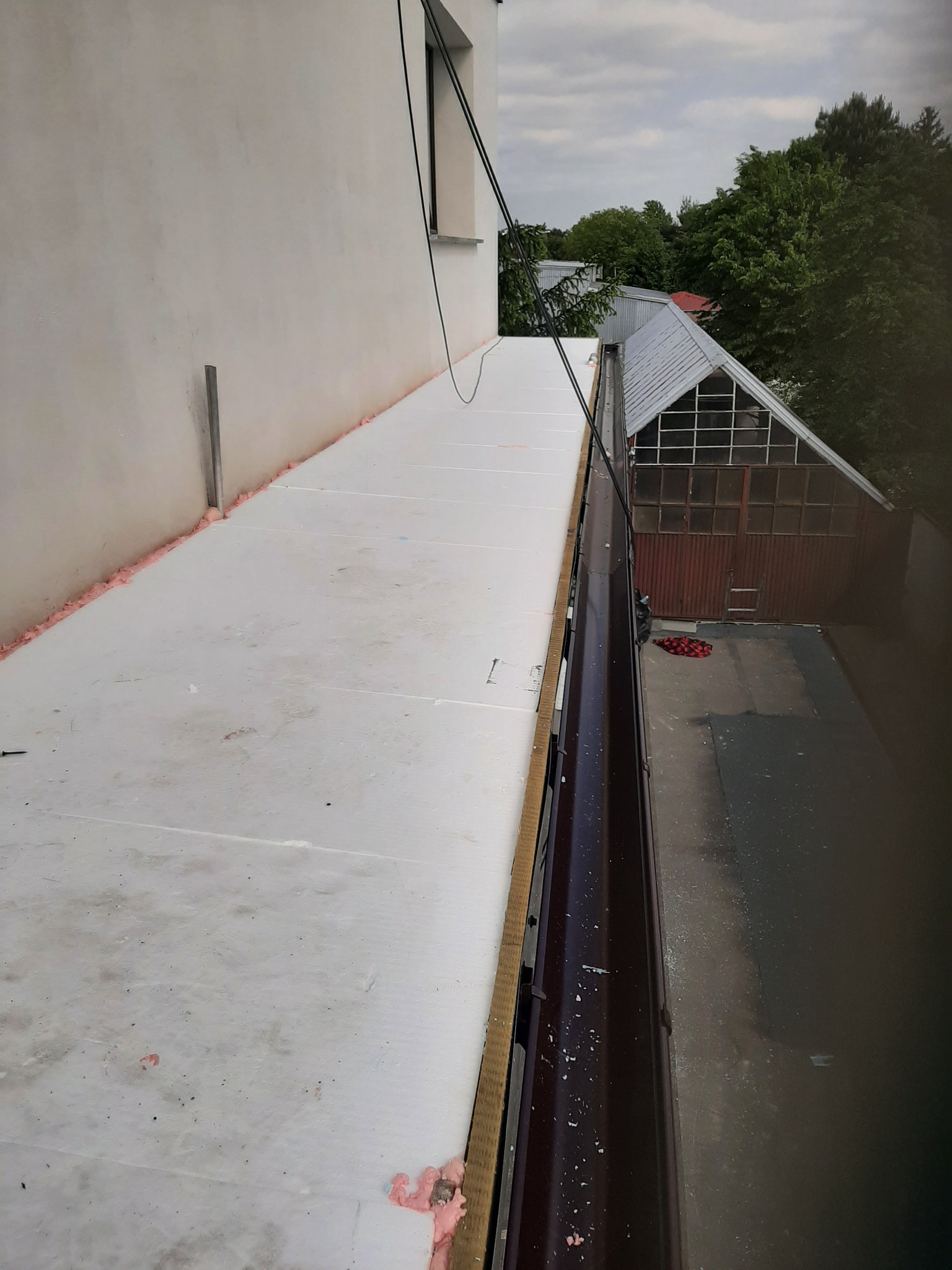 Białe płyty izolacyjne, prawdopodobnie styropian, ułożone na dachu budynku, widoczne rynny i fragment budynku gospodarczego w tle.