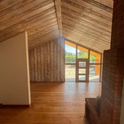 Wnętrze domu w trakcie budowy z drewnianym wykończeniem ścian, podłogi i sufitu, widoczne okno tarasowe z drewnianą ramą i fragment kominka z cegły.