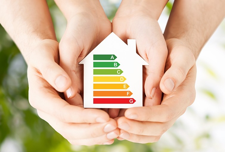 Dłonie trzymają biały model domu z kolorową skalą efektywności energetycznej od A do G na tle rozmytej zieleni.